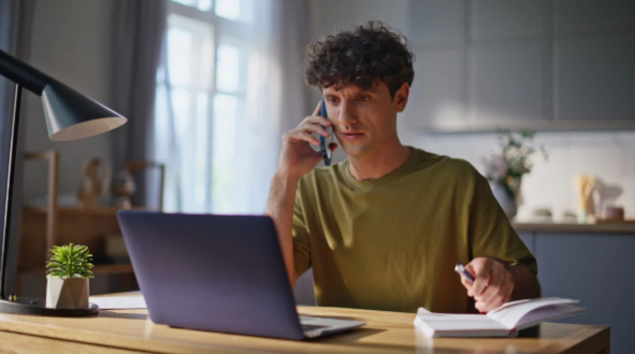Ein Mann sitzt am Küchentisch und telefoniert, während er auf seinen aufgeklappten Laptop blickt.
