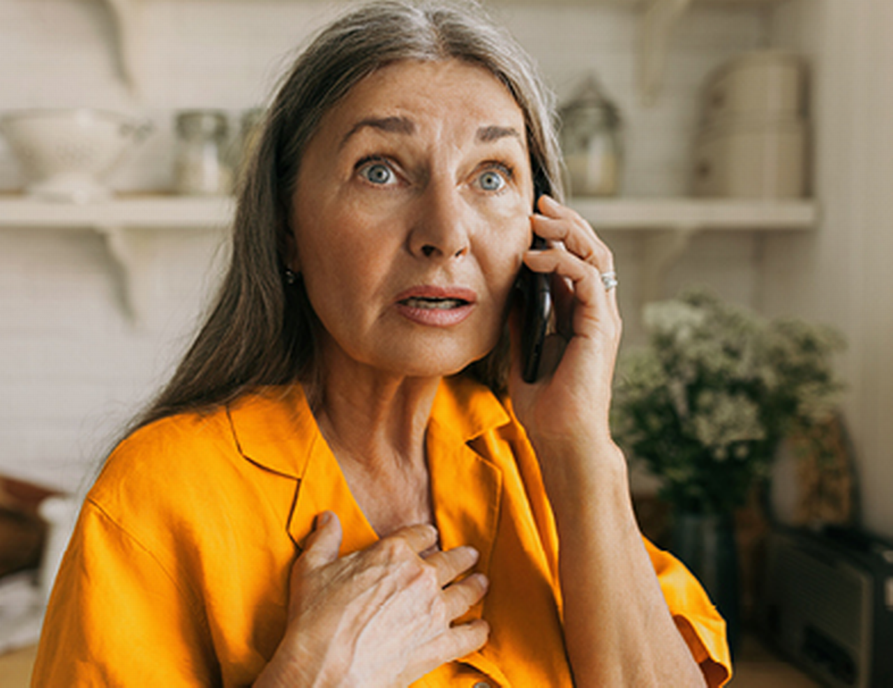 Frau mit schockiertem Gesichtsausdruck greift sich mit der Hand ans Herz, während sie sich ein Telefon ans Ohr hält.